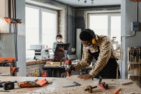 Two people working in a woodworking shop, using tools and wearing safety gear