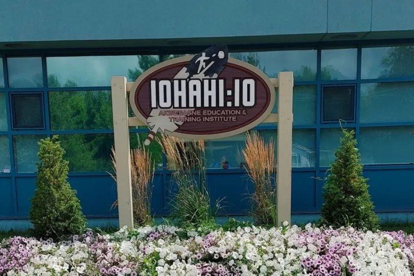 An outdoor sign for the Iohahi:io Akwesasne Education & Training Institute, featuring a large feather graphic, situated in front of a blue building and a garden of purple and white flowers