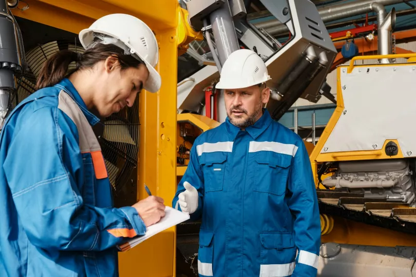 Two people in hardhats converse, one is writing something on a notepad