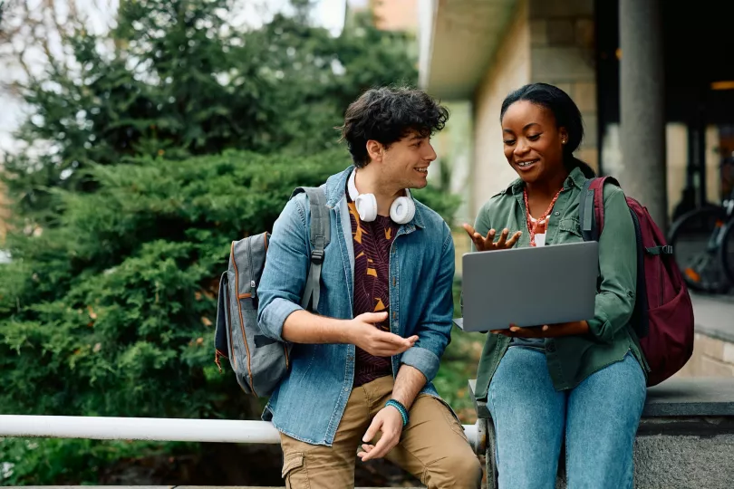 Two people converse in an academic setting; one holds a laptop