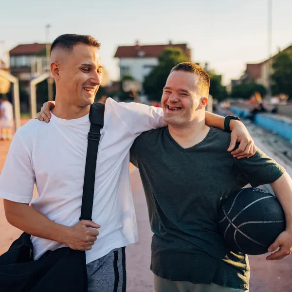Cheerful man with Down syndrome hugs his cheerful best friend as they walk on an outdoor sports court, ready to play basketball