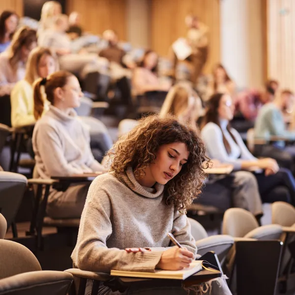 Female college student writing an exam during a class at lecture hall. Her classmate are in the background.