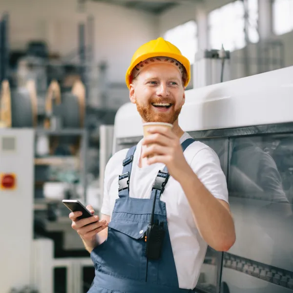 A person in a hardhat smiles and drinks a beverage in an industrial setting