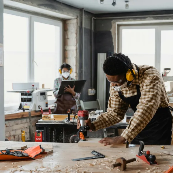 Two people working in a woodworking shop, using tools and wearing safety gear