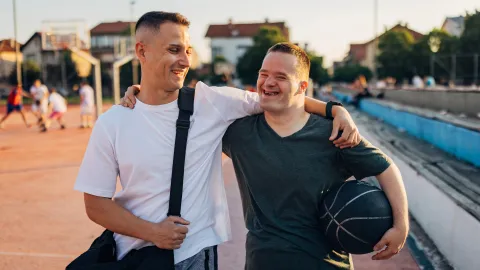 Cheerful man with Down syndrome hugs his cheerful best friend as they walk on an outdoor sports court, ready to play basketball
