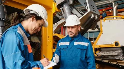 Two people in hardhats converse, one is writing something on a notepad