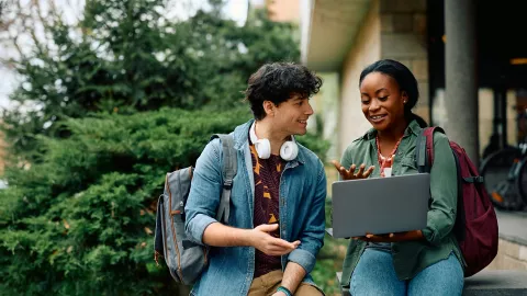 Two people converse in an academic setting; one holds a laptop