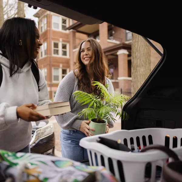 Two college students unloading belongings from a car trunk, excitedly moving into a new apartment and starting their independent lives