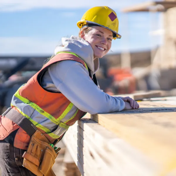 A female construction worker leans on a stack of lumber as she turns to pose for a portrait