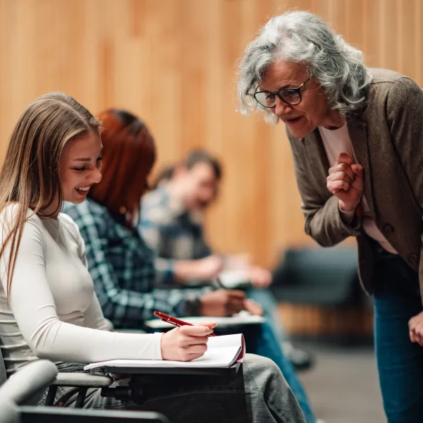 Senior female professor mentoring young adult student in a university classroom, providing guidance during a college lecture