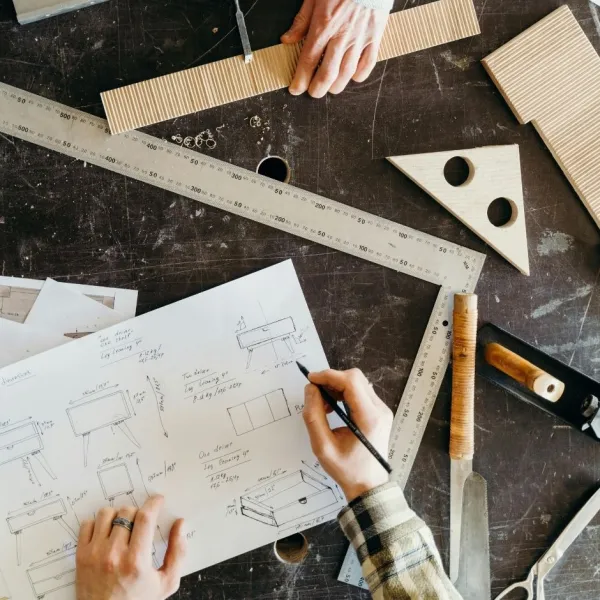 An overhead shot of two woodworkers at a dark workbench filled with tools, technical drawings, and a wooden furniture sketch.