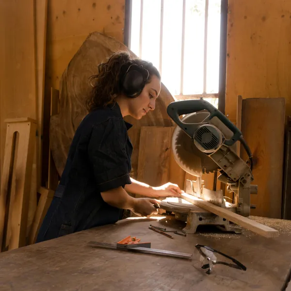 A woman wearing ear protection uses a miter saw to cut a piece of wood in a sunlit woodworking shop.