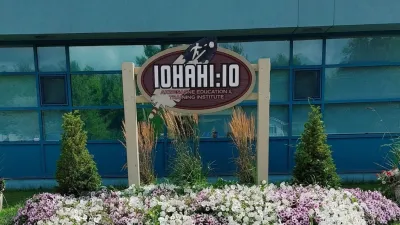 An outdoor sign for the Iohahi:io Akwesasne Education & Training Institute, featuring a large feather graphic, situated in front of a blue building and a garden of purple and white flowers