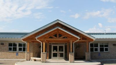 An exterior view of the Kenjgewin Teg entrance, showcasing a light-colored brick building with a large wood-beamed portico and a peaked roof