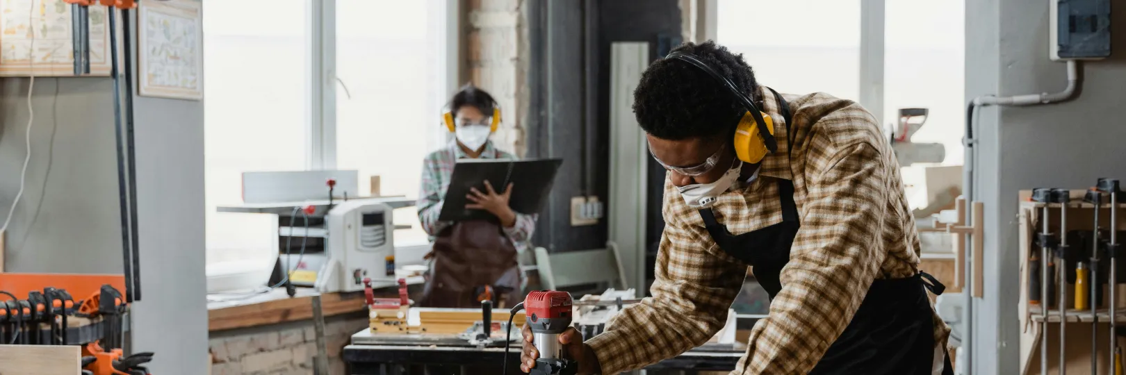 Two people working in a woodworking shop, using tools and wearing safety gear